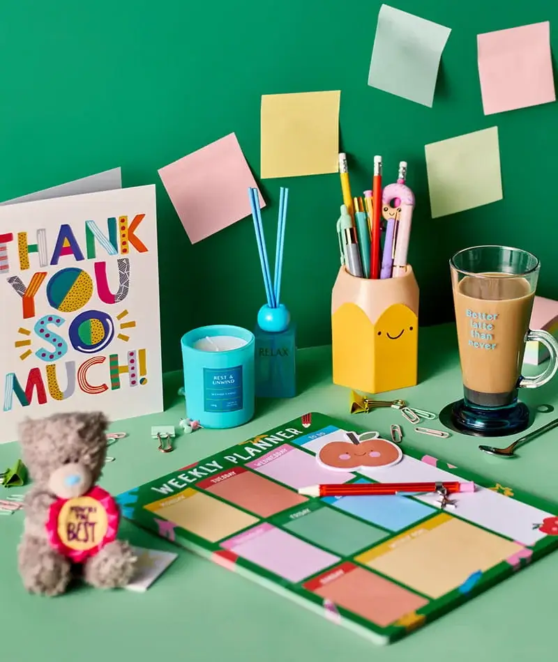 Colourful desk setup with thank you card, weekly planner, pens, sticky notes, candle, teddy bear, and latte on a green background